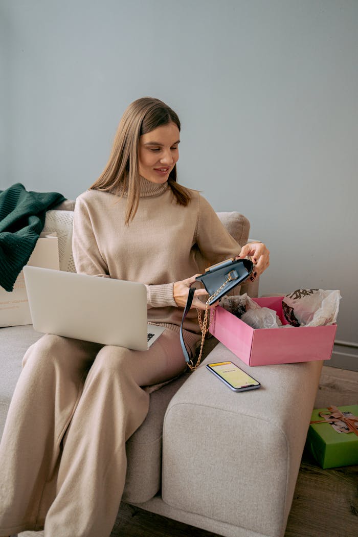 A woman unboxes a new handbag while online shopping at home using her laptop.